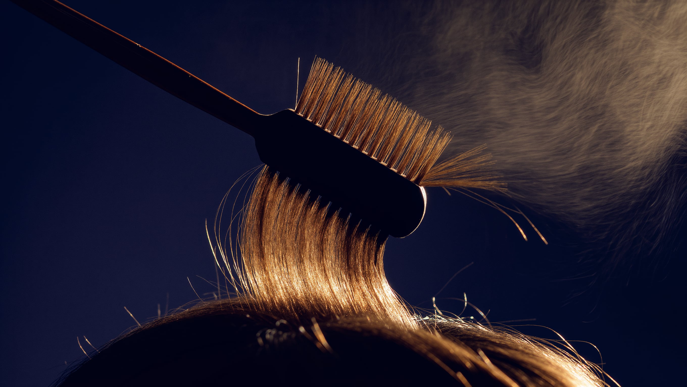 Rundbürste und eine gespannte Haarsträhne im warmen Föhnstrom, feine Lichtreflexe auf glatter Oberfläche